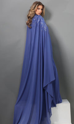 A woman with long wavy hair stands on a white platform against a gray background, showcasing timeless mother-of-the-bride grace in the Jovani 49742 long formal dress—a floor-length royal blue chiffon gown adorned with beaded embellishments and elegant embellished cape sleeves.