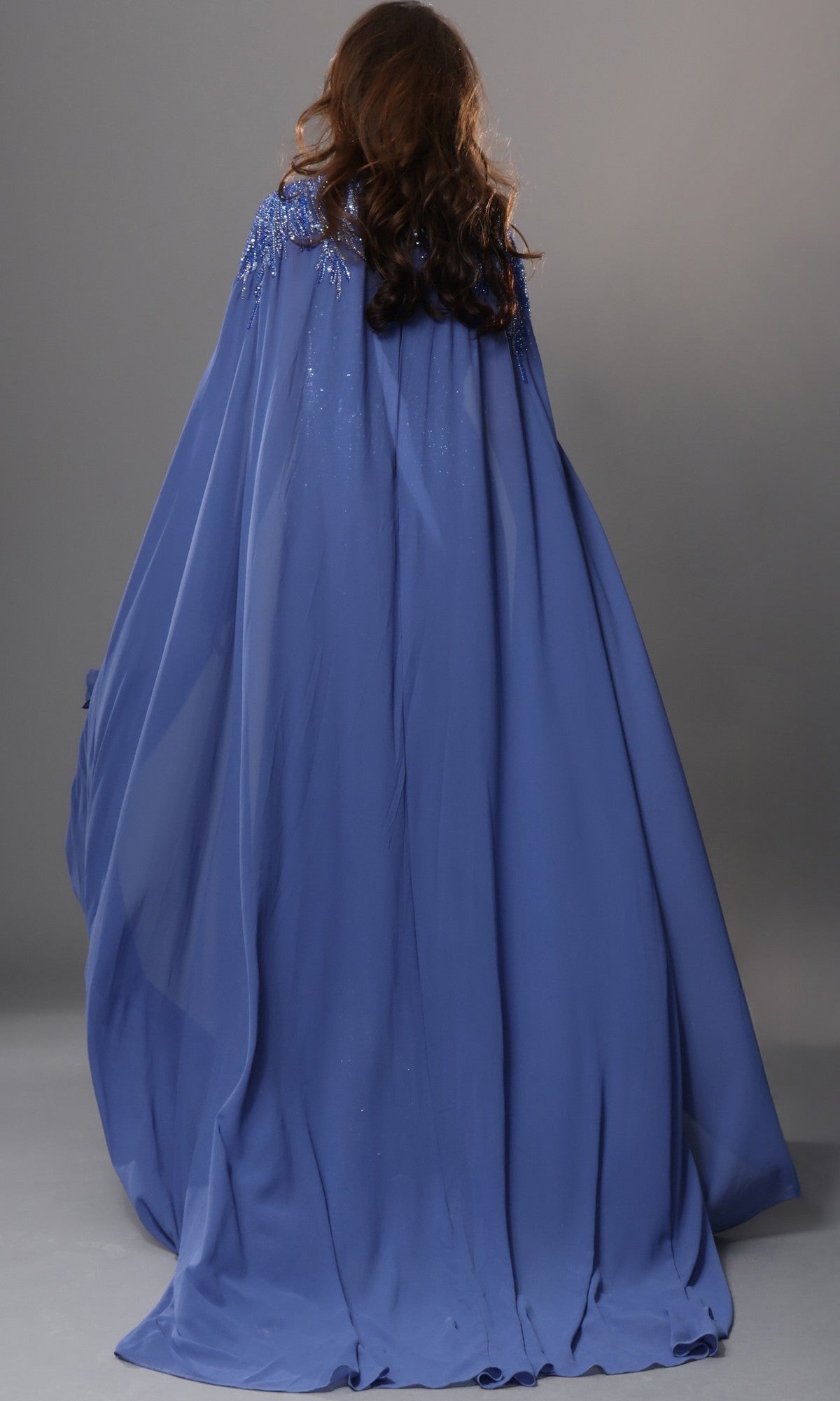 A person with long brown hair is shown from the back, wearing the Jovani 49742—a flowing, floor-length blue chiffon formal dress distinguished by embellished cape sleeves and intricate shoulder details—set against a plain gray background, making it an elegant choice for a mother-of-the-bride.