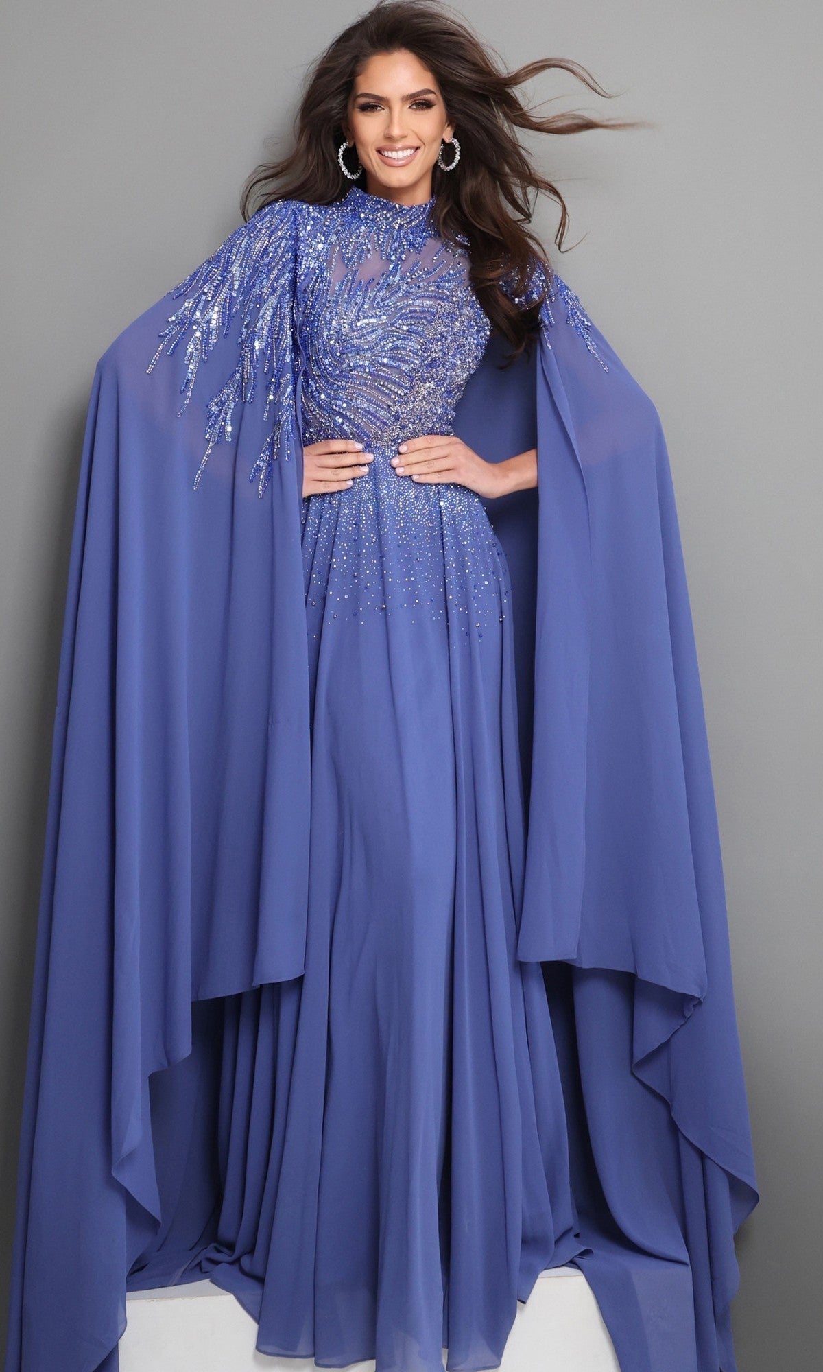 A woman stands smiling against a plain gray background, wearing the Jovani 49742—a floor-length, royal blue chiffon formal dress featuring sequin embellishments and dramatic embellished cape sleeves—an elegant choice perfect for a mother-of-the-bride.