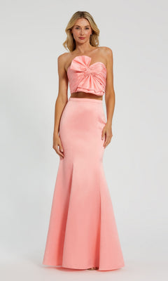 A woman wears the Mac Duggal 49832 Strapless Two-Piece Prom Gown in light pink, featuring a large bow on the strapless bodice and a graceful mermaid skirt, set against a plain backdrop.