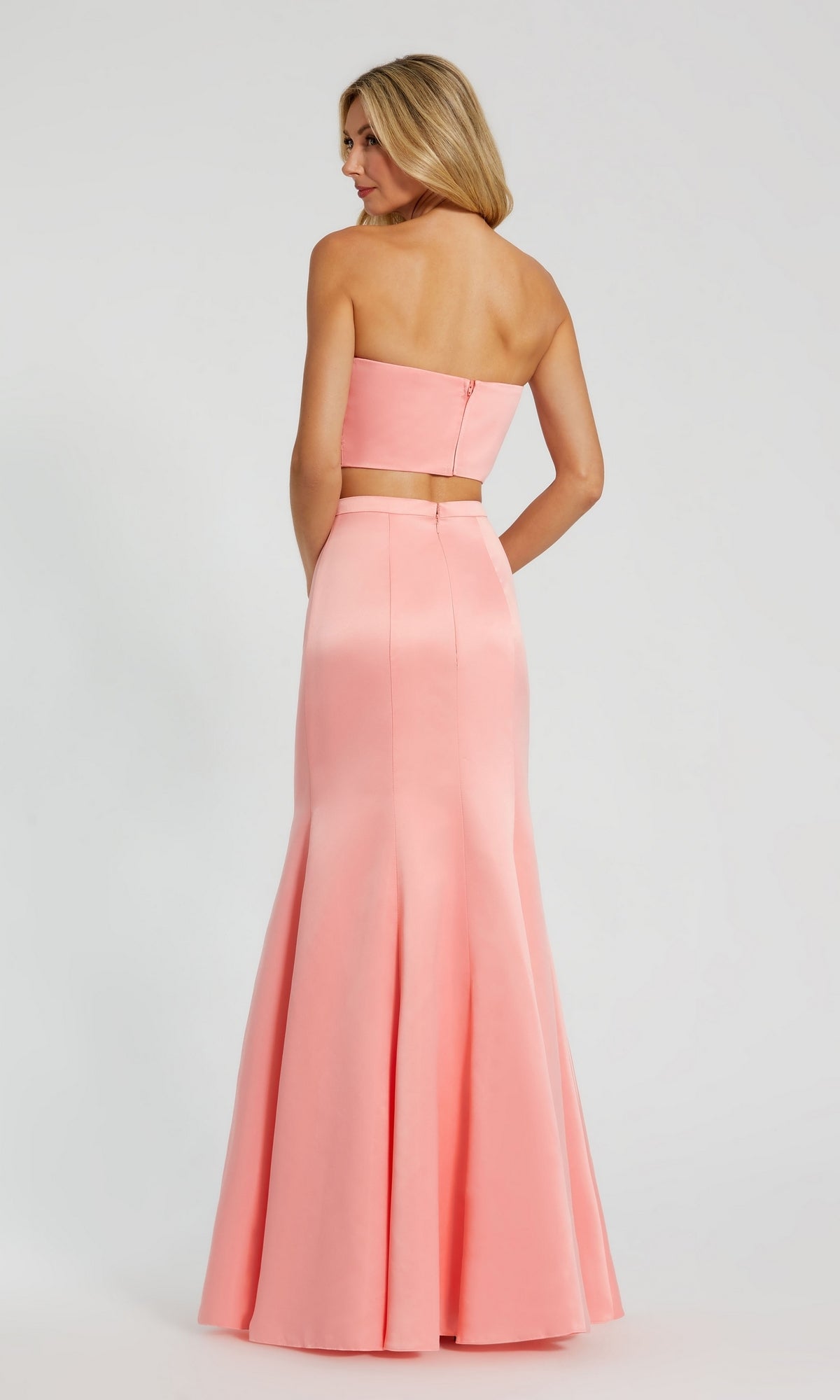 A woman showcases the Strapless Two-Piece Prom Gown: Mac Duggal 49832 in light pink, facing away to highlight its fitted bodice and flared mermaid skirt against a plain background.