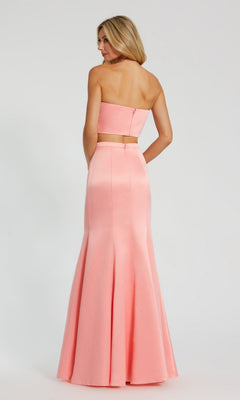 A woman showcases the Strapless Two-Piece Prom Gown: Mac Duggal 49832 in light pink, facing away to highlight its fitted bodice and flared mermaid skirt against a plain background.