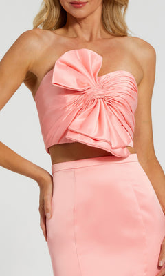 A woman models the Strapless Two-Piece Prom Gown: Mac Duggal 49832—a pale pink satin set with a strapless crop top featuring a gathered bow and a matching high-waisted mermaid skirt for an elegant prom look.