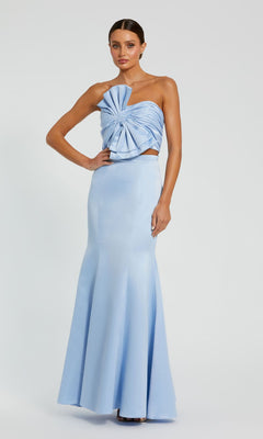 A woman models the Mac Duggal 49832, a strapless two-piece prom gown in light blue, featuring a dramatic sculpted bow on the bodice and a floor-length silhouette, standing against a plain background.