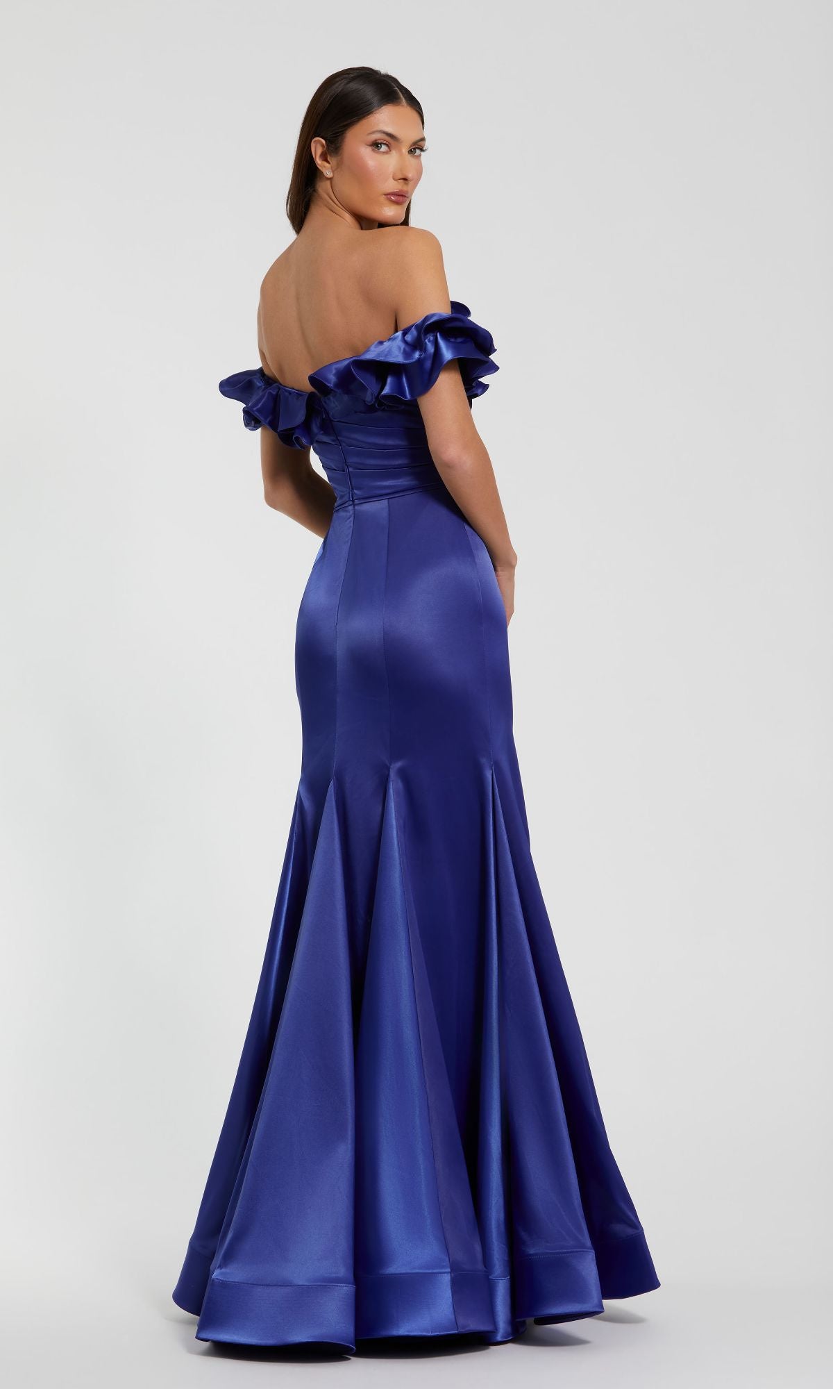 A woman stands against a plain background, looking over her shoulder in the Mac Duggal 49834 Ruffled Long Satin Prom Dress, a royal blue off-the-shoulder gown with floor-length ruffled sleeves.