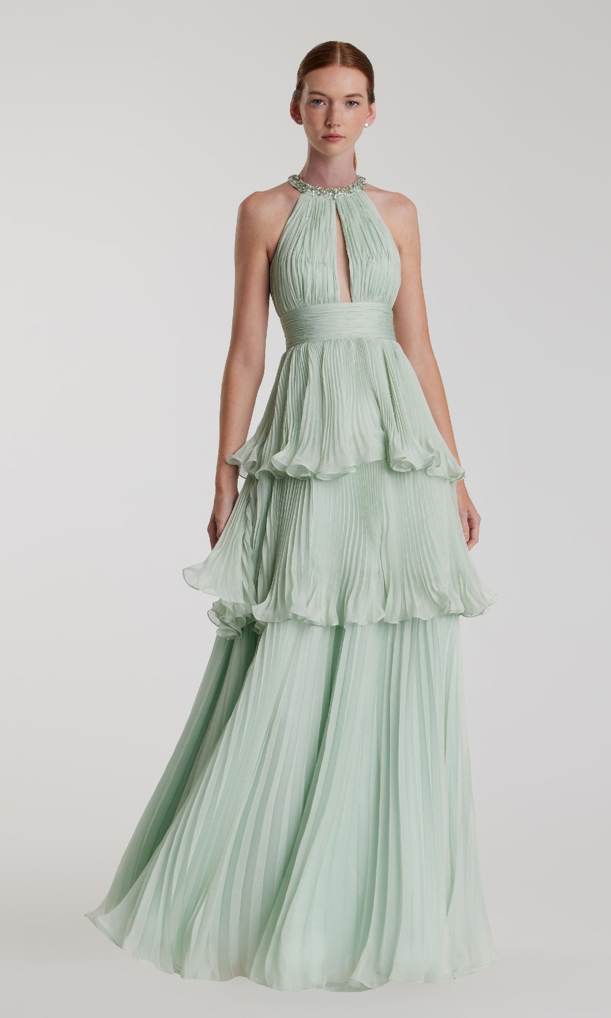 A woman wears the Mac Duggal 49835—an elegant long mint green formal dress with pleated, tiered layers and a halter-neck keyhole neckline—standing against a plain background.