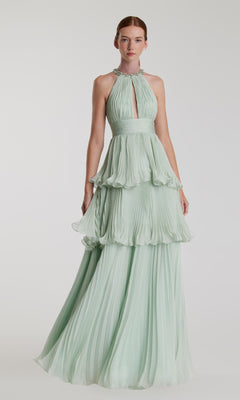 A woman wears the Mac Duggal 49835—an elegant long mint green formal dress with pleated, tiered layers and a halter-neck keyhole neckline—standing against a plain background.