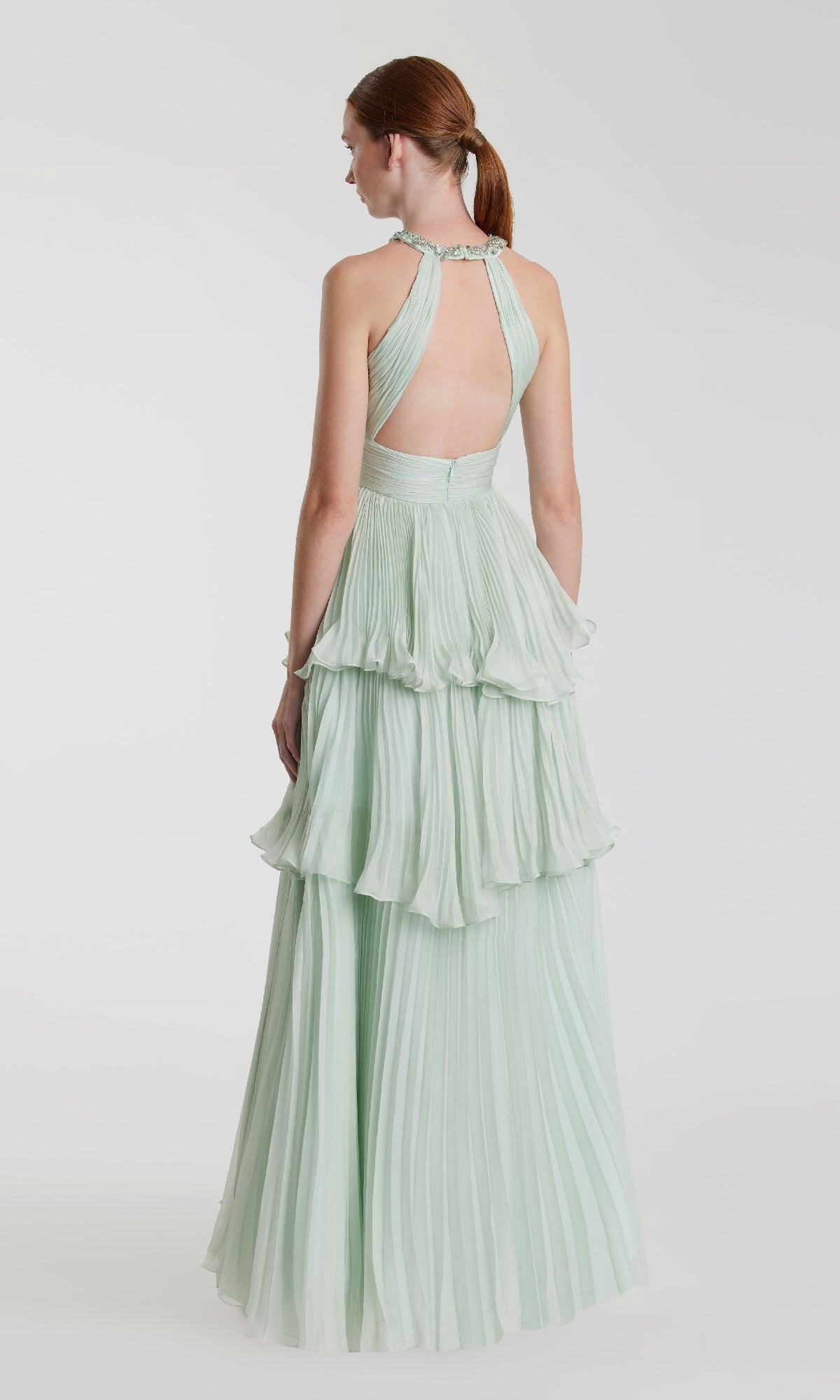 A red-haired woman in a low ponytail wears the Mac Duggal 49835 long mint green formal dress, featuring a backless halter-neck and tiered pleats, as she stands against a plain white background.