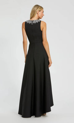 A woman stands facing away, wearing the Mac Duggal 49844 High-Low Formal Dress—a sleeveless black crepe gown with white embellishments on the upper back and an elegant a-line skirt.