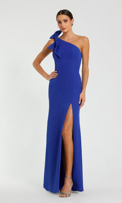 A woman stands against a plain light background wearing the Mac Duggal 49862 one-shoulder long formal dress in royal blue, featuring a side bow and thigh-high slit.