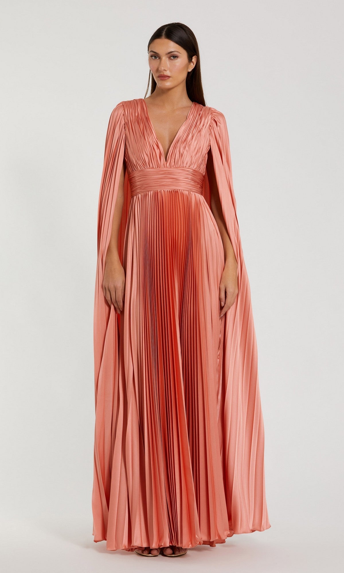 A woman stands against a plain background wearing the Mac Duggal 49872 Cape-Sleeve Long Formal Dress, a floor-length peach gown with pleats, cape-style sleeves, and a deep V-neckline.