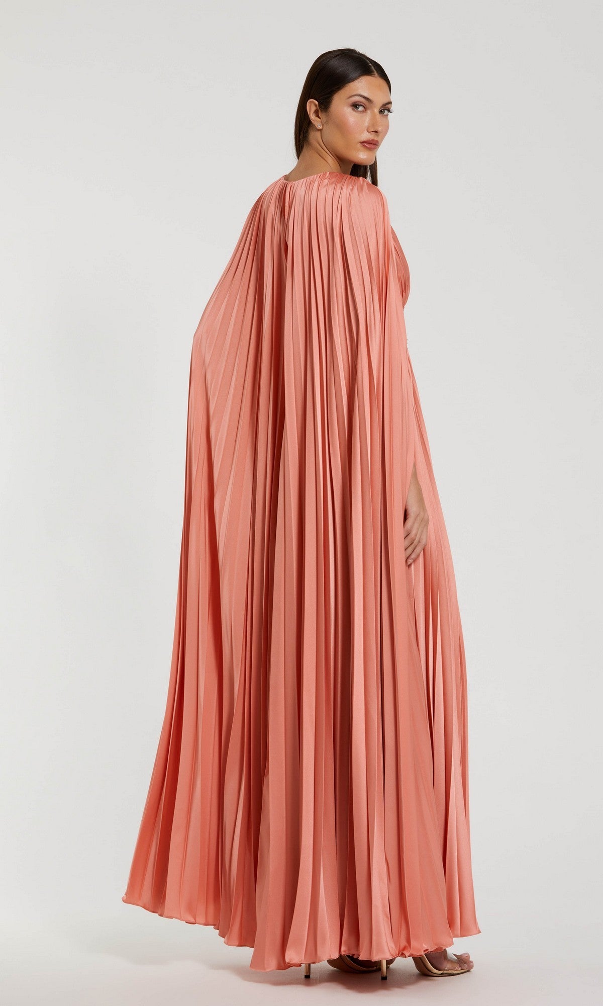 A woman stands against a plain light background wearing the Mac Duggal 49872 Cape-Sleeve Long Formal Dress in a peach hue, featuring elegant pleats and a flowing cape-like design.