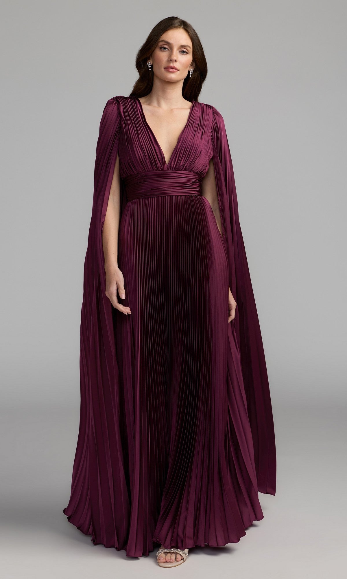 A woman models the Mac Duggal 49872 Cape-Sleeve Long Formal Dress in deep burgundy, featuring pleats, a floor-length silhouette, deep V-neckline, and cape sleeves against a plain gray background.