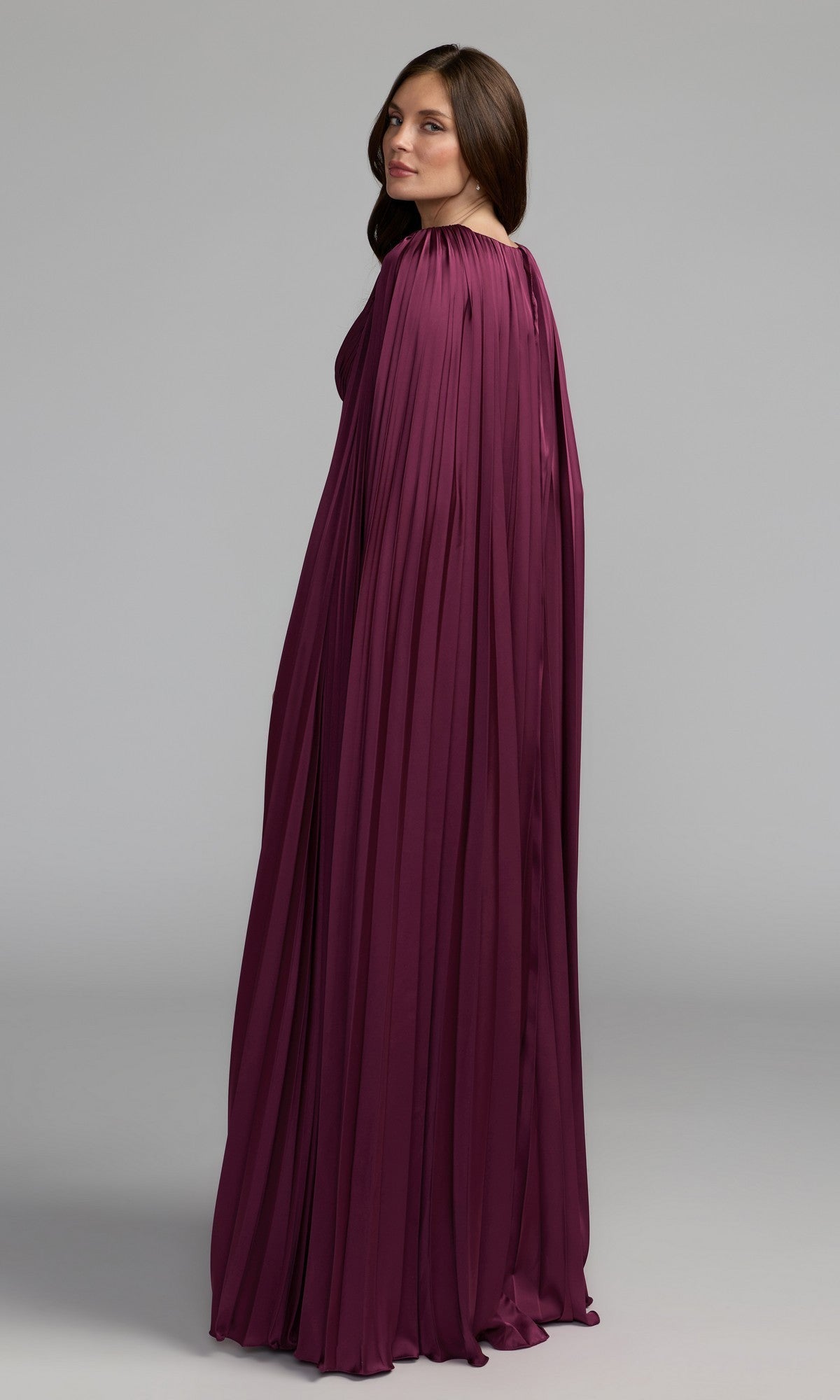 A woman in profile wears the Mac Duggal 49872 Cape-Sleeve Long Formal Dress in deep purple, featuring pleats and a cape-style design, set against a plain light gray background.