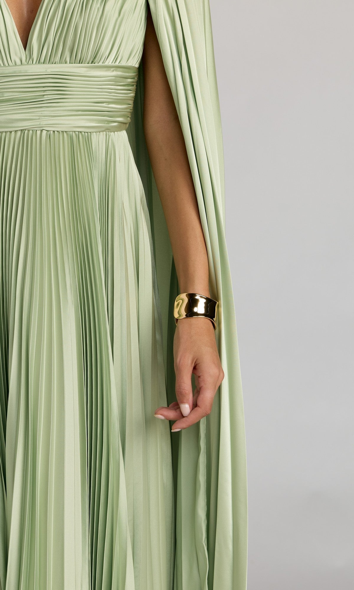 A person wearing the Mac Duggal 49872 Cape-Sleeve Long Formal Dress in light green, featuring pleats, a wide waistband, and paired with a gold cuff bracelet.
