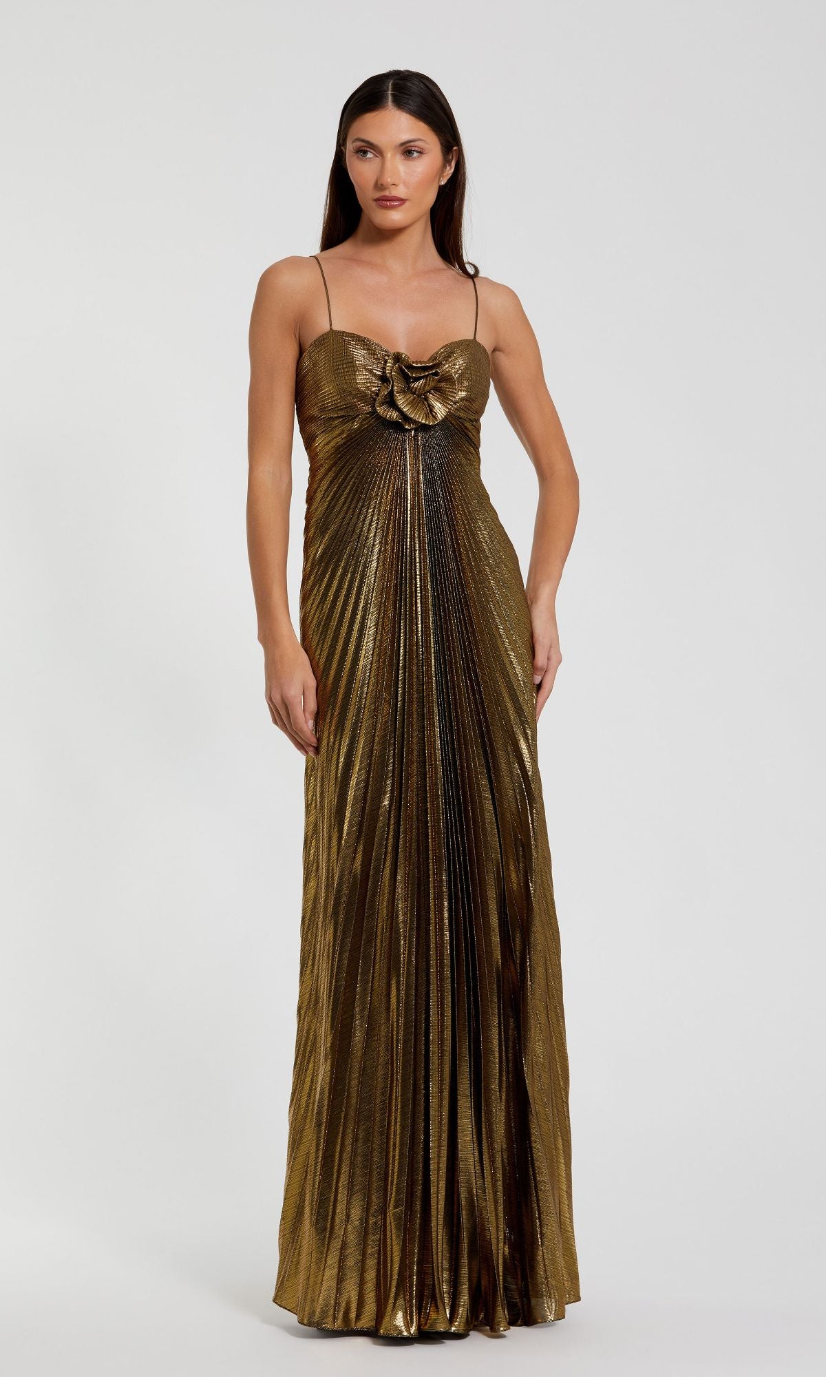 A woman stands against a plain background wearing the Mac Duggal 49905 Metallic Gold Long Prom Dress, featuring thin straps, pleated fabric, and a large decorative bow on the bodice.