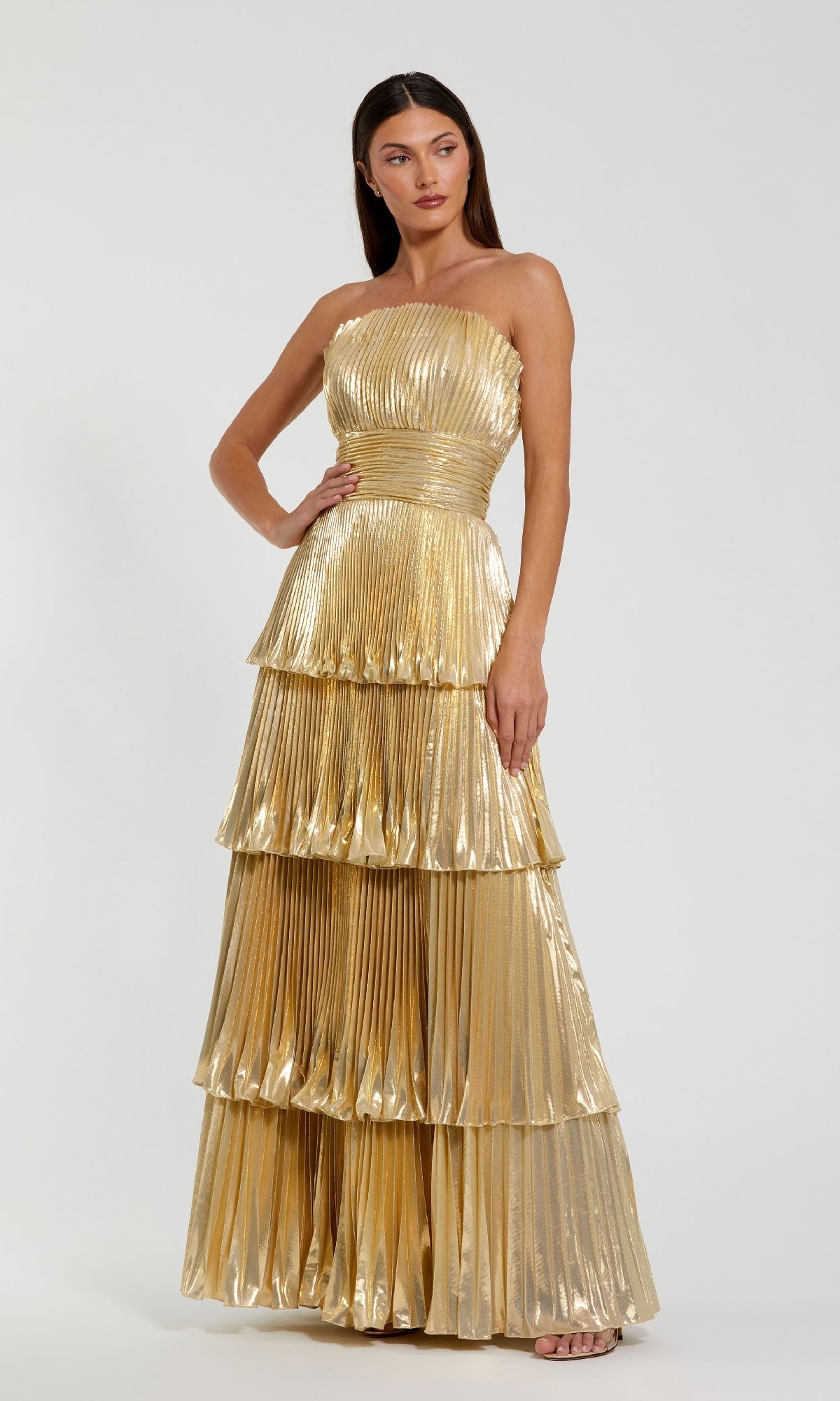 A woman models the Mac Duggal 49911 long formal dress—a strapless gold gown with pleated, tiered layers and a metallic sheen—standing against a plain light background.
