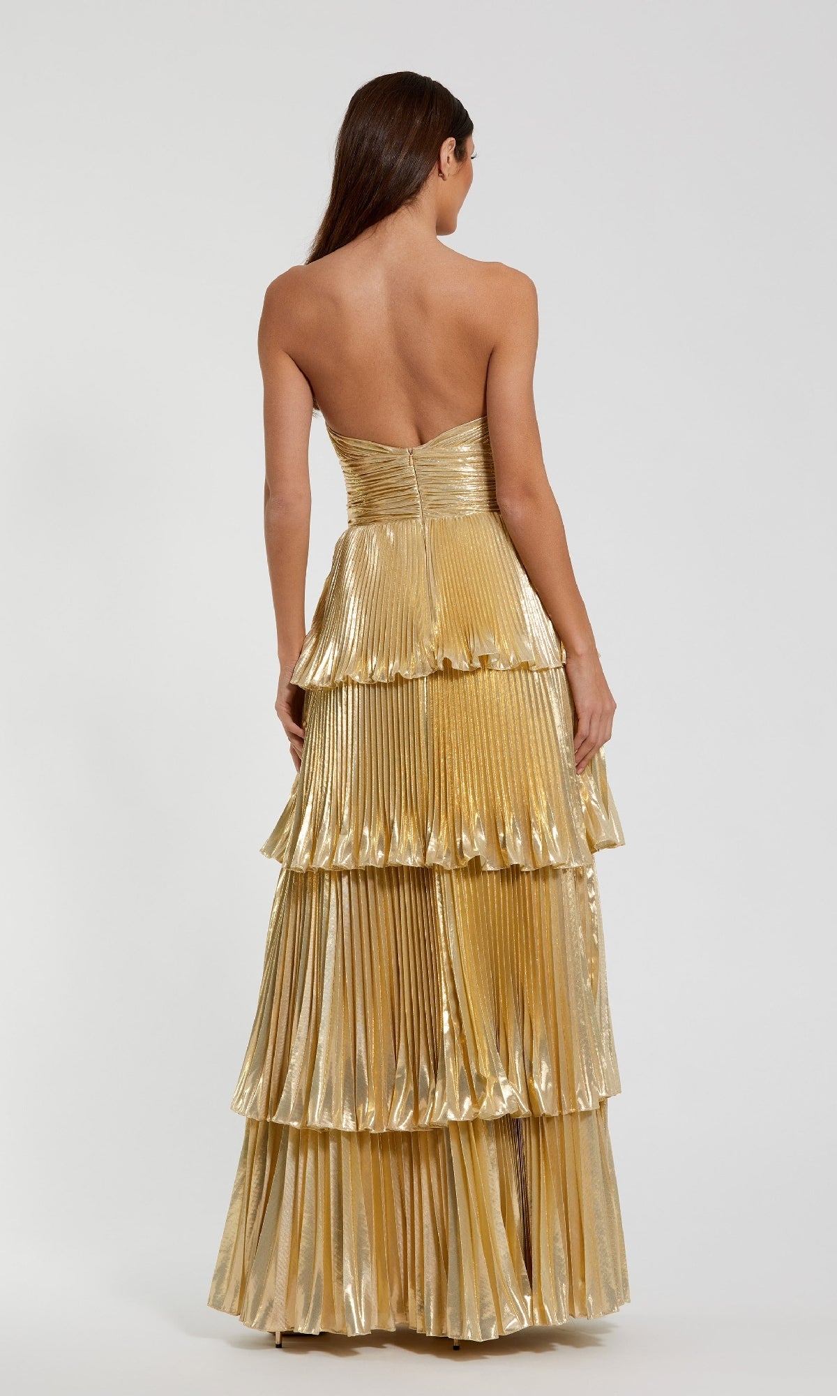 A woman with long brown hair stands with her back to the camera in the Mac Duggal 49911 long formal dress, a strapless gold gown with pleated, tiered layers, set against a plain background.