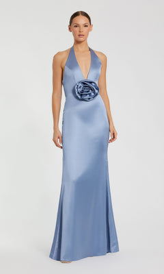 A woman stands against a white background, wearing the Mac Duggal 49913 Open-Back Long Halter Prom Dress—a floor-length, light blue satin gown featuring a halter neckline and a large rose detail at the waist.