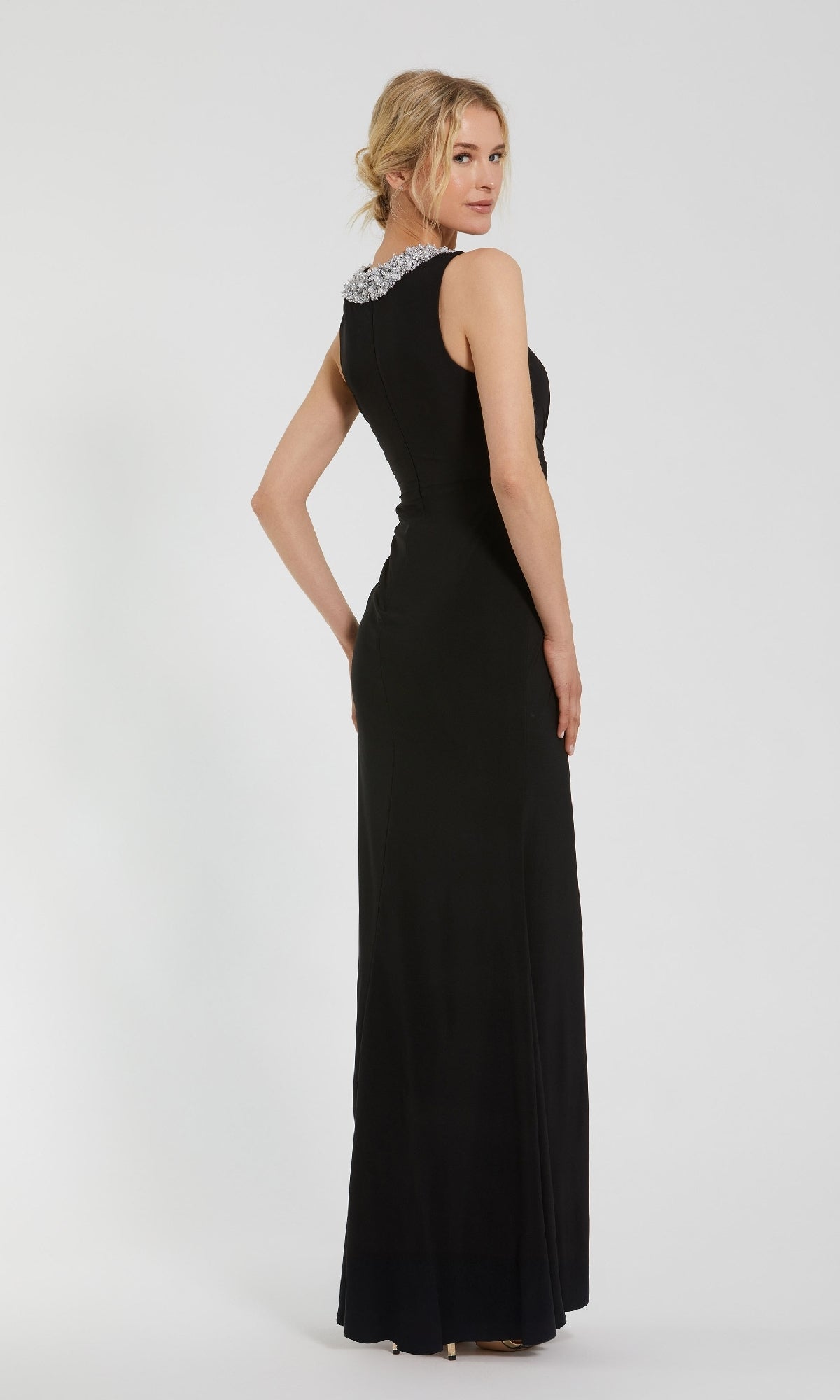 A woman in the Jeweled Long Black Formal Dress (Mac Duggal 49952), a sleeveless, floor-length gown with a decorative collar, stands turned slightly away from the camera.