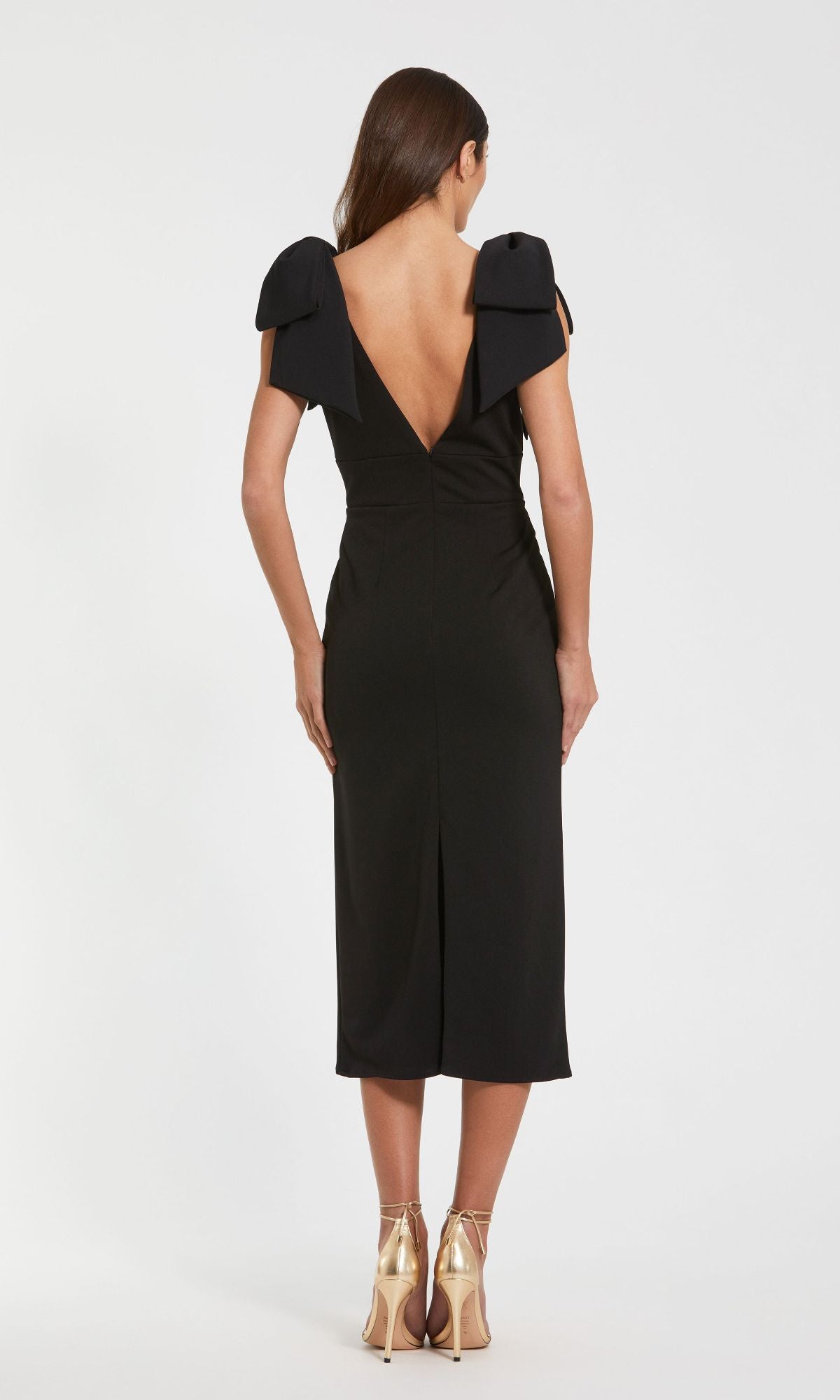 A woman faces away from the camera in a Mac Duggal 49954 V-Neck Midi Wedding Guest Dress, featuring a deep V-back and bow shoulder details, styled with gold heels against a plain backdrop.