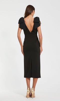 A woman faces away from the camera in a Mac Duggal 49954 V-Neck Midi Wedding Guest Dress, featuring a deep V-back and bow shoulder details, styled with gold heels against a plain backdrop.