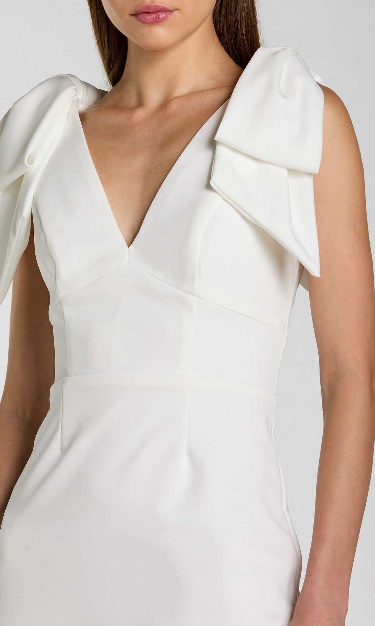 A woman wears the Mac Duggal 49954 V-Neck Midi Wedding Guest Dress, featuring a sleeveless white design, deep V-neck, large bow-inspired shoulder accents, and elegant tea-length style.