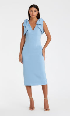 A woman wears the Mac Duggal 49954 V-Neck Midi Wedding Guest Dress, sleeveless and light blue with large shoulder bows, paired with strappy high-heeled sandals, standing against a plain white background.