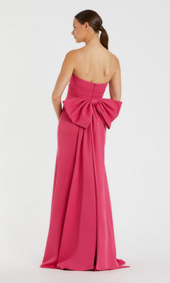 A woman wears the Mac Duggal 49967 long formal dress—a pink strapless gown featuring an oversized bow on the back—standing against a plain white background.
