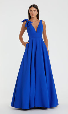 A woman in the Mac Duggal 49972 Shoulder-Bow Long Formal Dress, featuring a sleeveless royal blue design with a deep V-neck and large shoulder bow, stands against a plain light background.