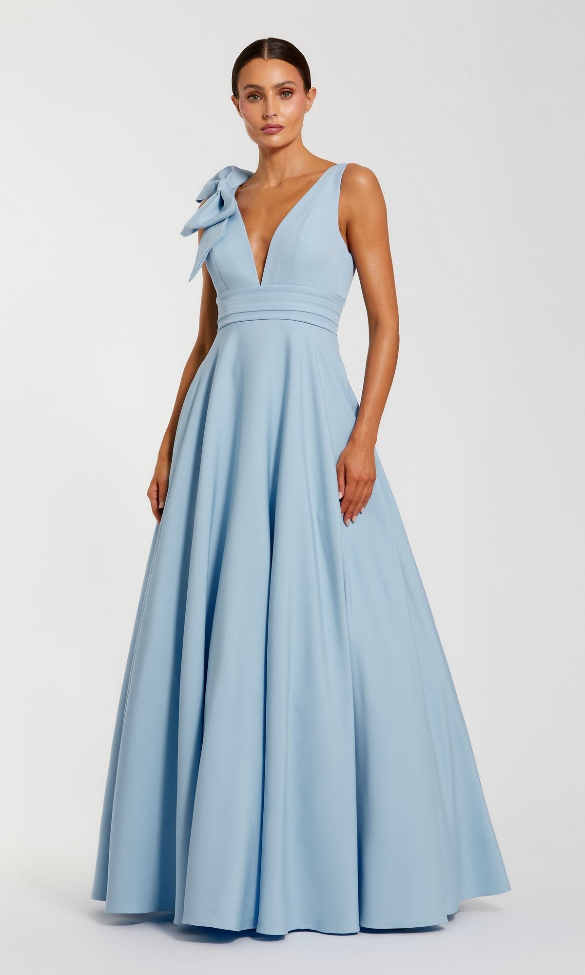 A woman stands against a plain background wearing the Mac Duggal 49972 Shoulder-Bow Long Formal Dress, a sleeveless, floor-length light blue gown with a deep V-neckline and an oversized bow on one shoulder.