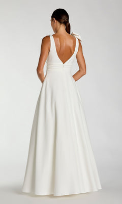 A woman in the Mac Duggal 49972 Shoulder-Bow Long Formal Dress, a sleeveless white gown with a V-back and full skirt, stands facing away with her hair tied back in a low ponytail.