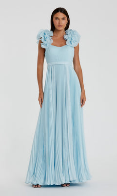 A woman stands facing the camera against a plain background, wearing the Mac Duggal 49979 long formal dress in powder blue, featuring floor-length pleated organza and prominent ruffled shoulder accents.
