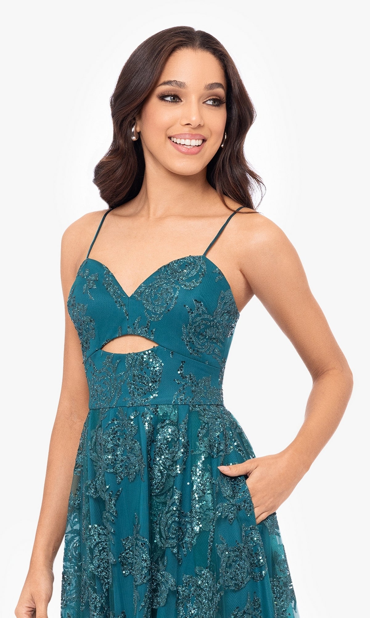 Smiling and looking to the side, a woman wears the Cut-Out Short Teal Glitter-Print Hoco Dress 4997BN—sleeveless with thin straps, floral glitter pattern, short hem, midriff cut-out, and pockets.