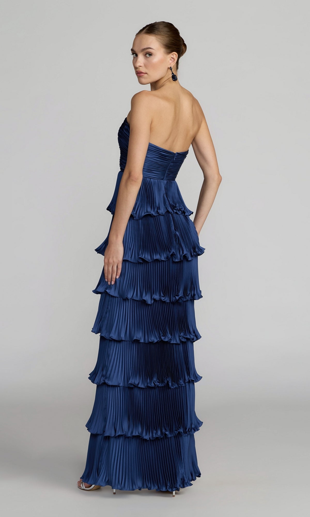 A woman poses against a plain background, looking over her shoulder in the Mac Duggal 49981 Strapless Pleated Long Formal Dress—a floor-length, navy blue gown with tiered pleats—making it a perfect choice for any special occasion.