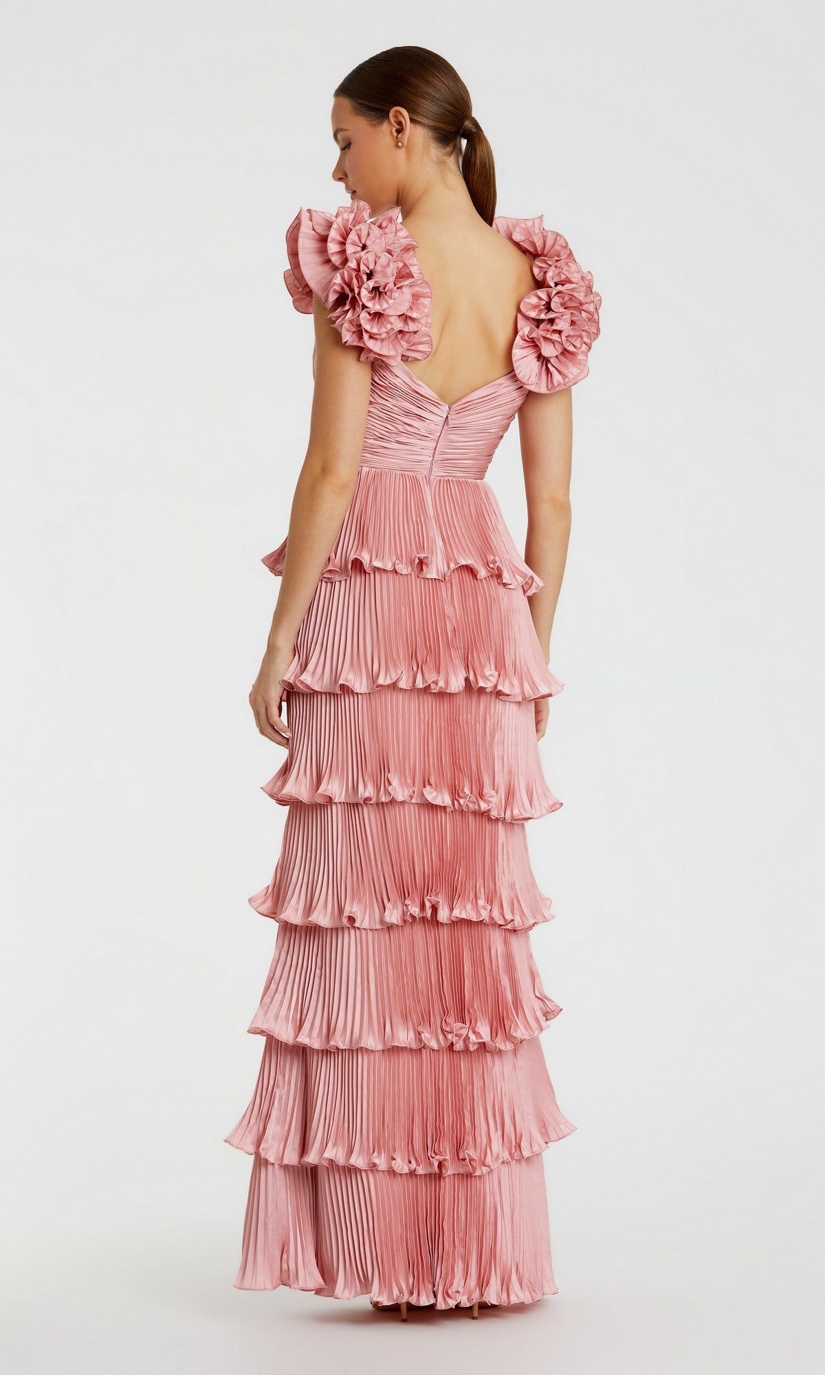 A woman in the Mac Duggal 49983 long formal dress—a floor-length pink pleated gown with ruffle tiers and bold floral shoulder details—stands with her back to the camera.