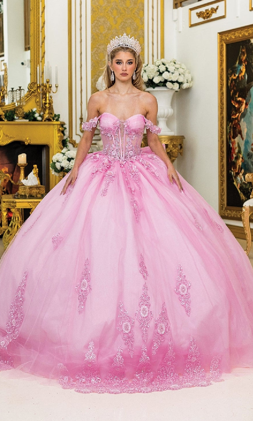 A woman wearing a jeweled crown and the Quinceanera Dress 5000 By Dancing Queen, featuring off-the-shoulder floral lace embroidery, stands in an elaborately decorated room.