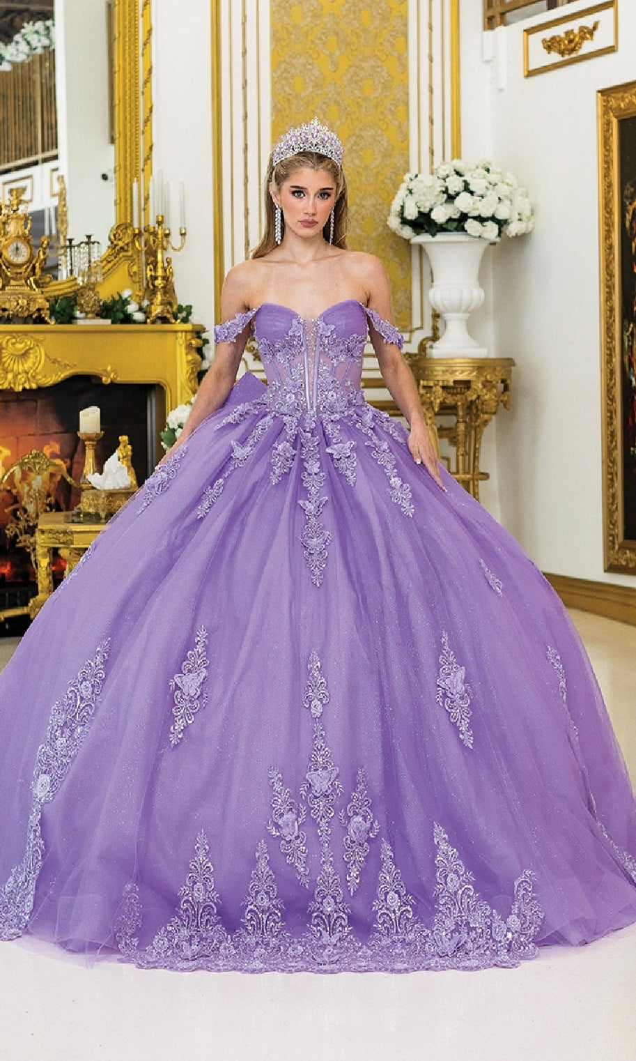 A woman in the Quinceanera Dress 5000 By Dancing Queen, featuring glitter tulle and floral lace embroidery, stands in a lavish gold room with a jeweled tiara.