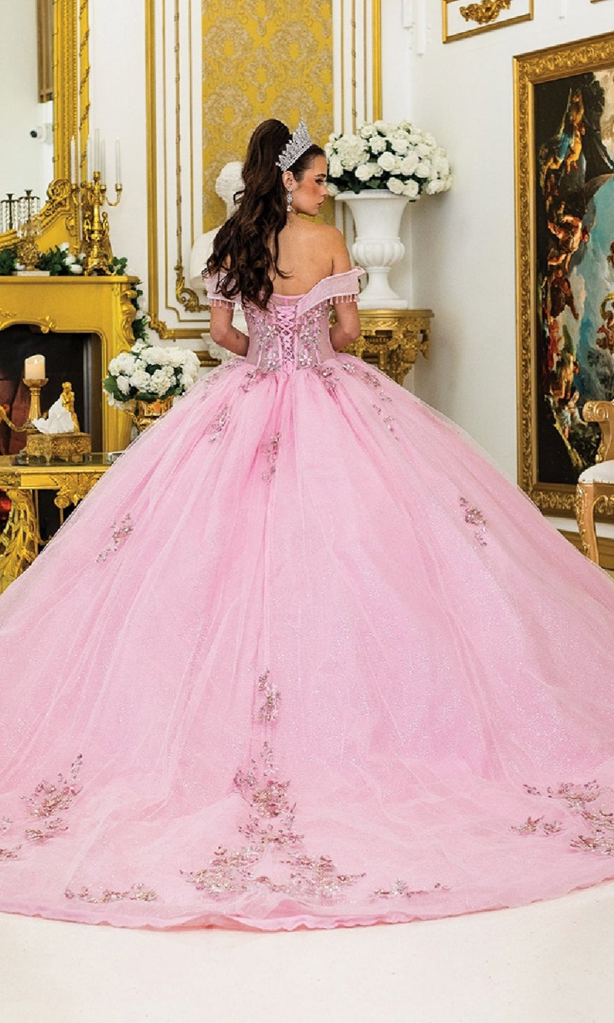 A woman wearing the Pink Quinceanera Dress 5001 By Dancing Queen, featuring a light pink glitter ball gown skirt and floral details, stands facing away in an ornate room with gold accents and white flowers.