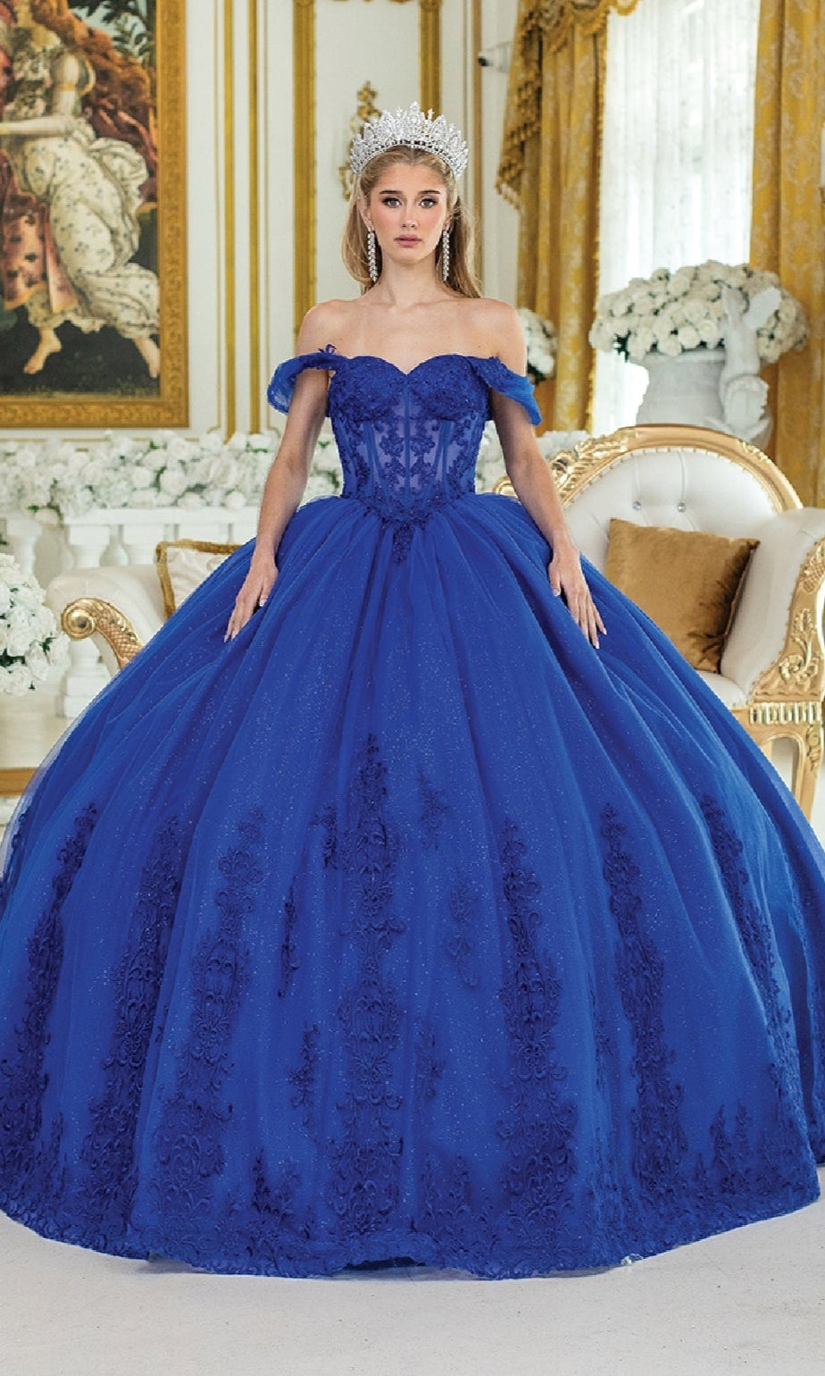 A woman in the Royal Blue Quinceanera Dress 5003 by Dancing Queen, featuring floral embroidery and a silver tiara, stands in an ornate room with gold accents and white floral arrangements.