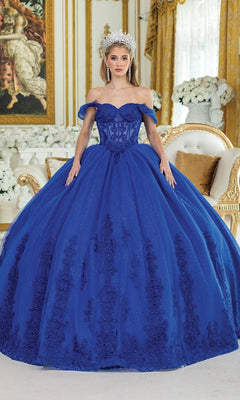 A woman in the Royal Blue Quinceanera Dress 5003 by Dancing Queen, featuring floral embroidery and a silver tiara, stands in an ornate room with gold accents and white floral arrangements.