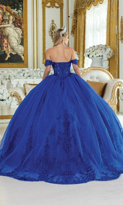 A woman in the Royal Blue Quinceanera Dress 5003 by Dancing Queen, featuring floral embroidery and a tiara, stands facing away in an ornate room with gold accents, white flowers, and a classical painting.