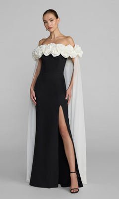 A woman wearing the Mac Duggal 50041 long formal dress in Black Ivory—a striking off-shoulder crepe gown with white floral details, a sheer white cape, a thigh-high slit, and paired with black heeled sandals.