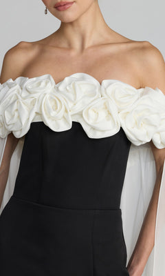 A woman wearing the Mac Duggal 50041 long formal dress, an off-the-shoulder black gown adorned with large white fabric roses along the neckline and sheer white draping on the arms.