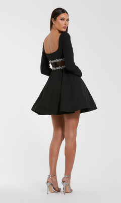 A woman wearing the Mac Duggal 50047 short party dress, a black fit-and-flare style with silver embellishments, stands in silver high heels, facing backward while turning her head toward the camera.