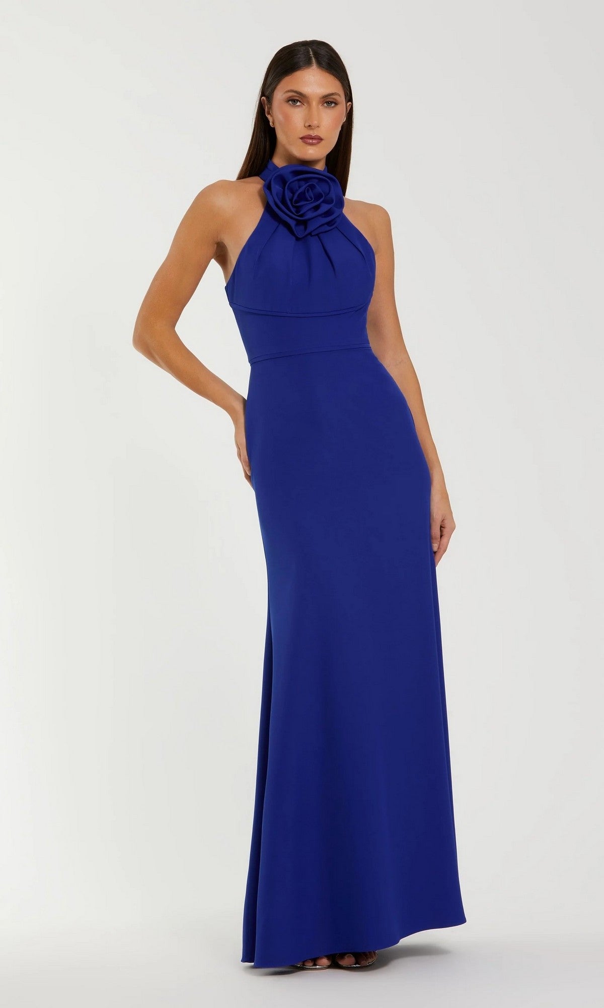 Woman in a sleeveless Mac Duggal 50053 long formal dress in cobalt, featuring a high halter neck and bold 3D rosette detail, stands against a plain light background.