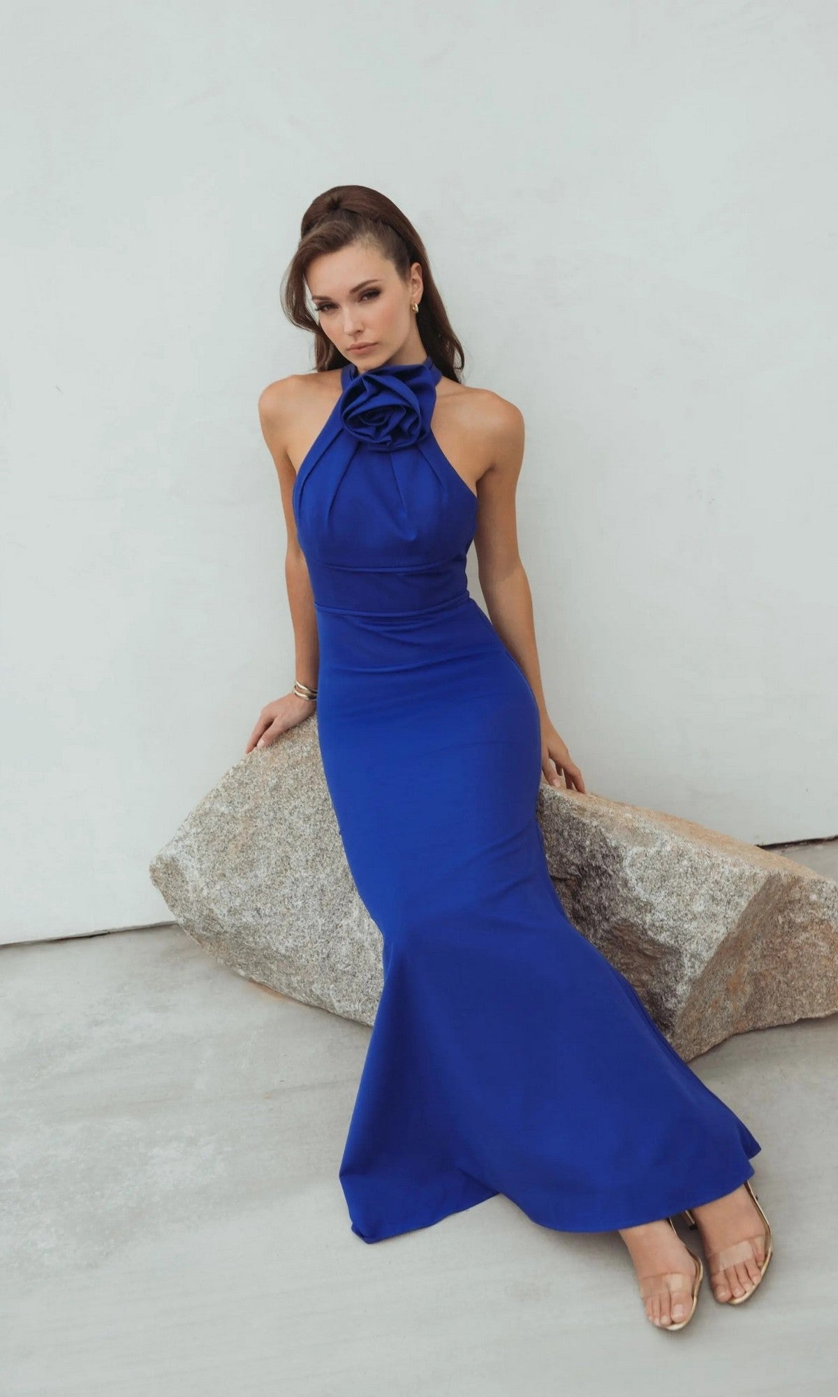 Woman wearing the Mac Duggal 50053 long formal dress in sleeveless royal blue with a 3D rosette neckline sits on a large rock against a light-colored background.