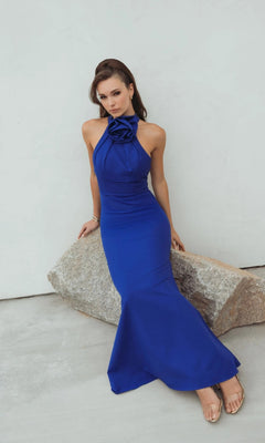 Woman wearing the Mac Duggal 50053 long formal dress in sleeveless royal blue with a 3D rosette neckline sits on a large rock against a light-colored background.