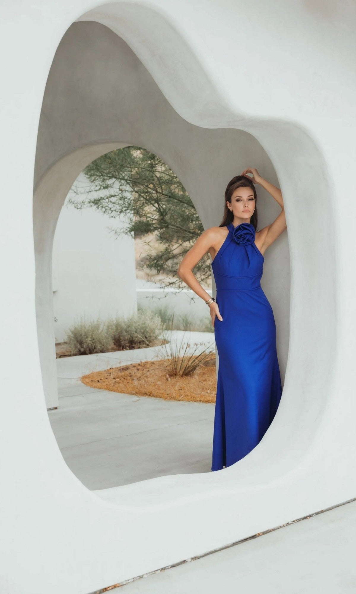 A woman in a sleeveless royal blue Long Formal Dress: Mac Duggal 50053 leans against an abstract white structure featuring curved openings.