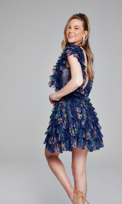 A woman stands sideways, smiling, in the Jovani Ruffled Short Print Homecoming Dress 50069—navy blue with floral print and ruffles—paired with nude heels, set against a plain gray background.
